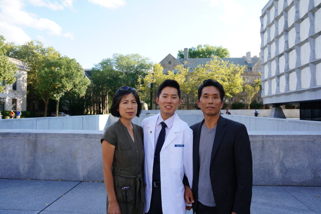 Photograph of three people, the man in the middle is in his 20s and has heritage from South Korea. He is wearing a doctor's white coat, button up shirt and tie. There is a man on his left and a woman on his left, they are both middle-aged. 