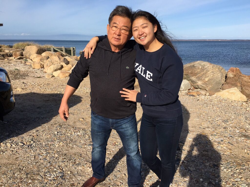 Photograph of a woman in her 20s who has heritage from Uzbekistan with light skin tone and long black hair, she is wearing a navy Yale crewneck sweatshirt and black jeans. She is side hugging a man next to her. He is in his 50s with light skin tone and black hair. They are standing on gravel and a body of water can be seen behind them.