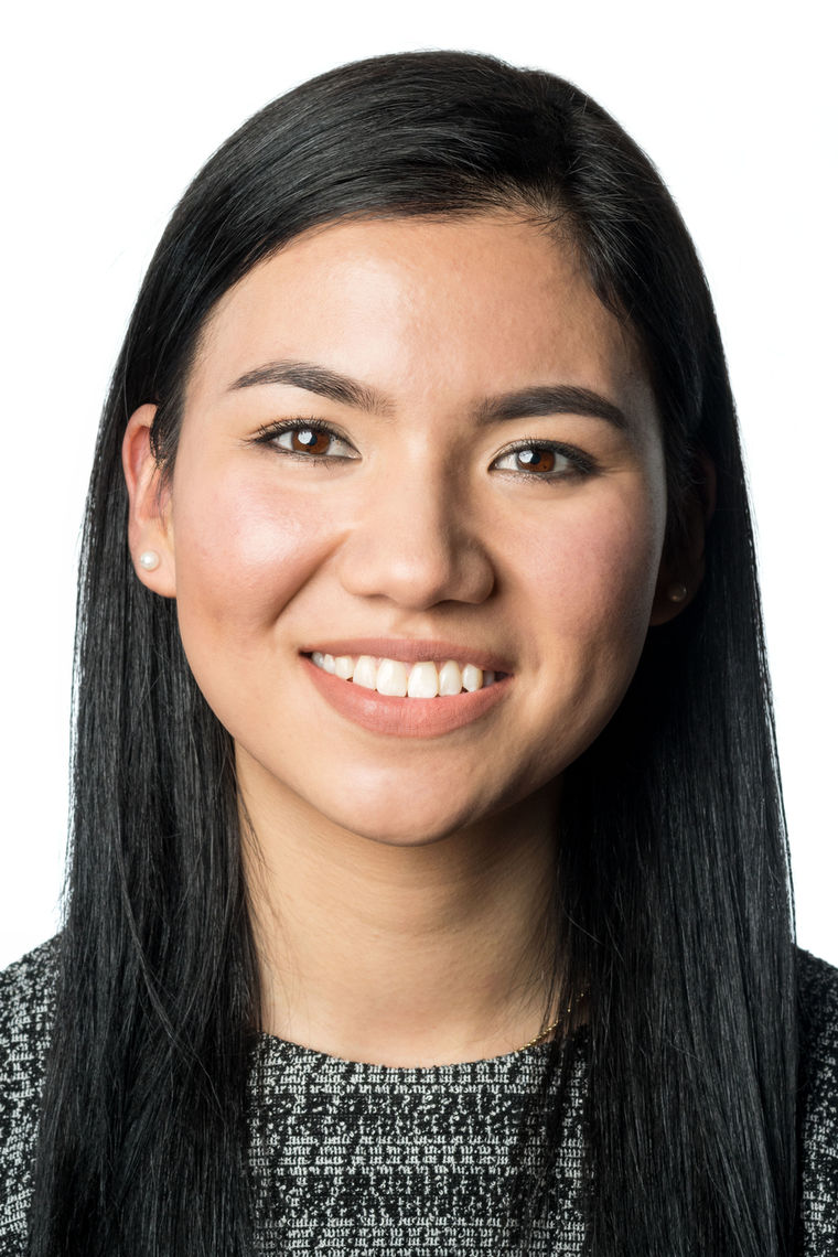 Headshot of a woman in her 20s who has heritage from Pakistan with light skin tone and long black hair parted on the side; she is wearing a black and white patterned high-neck top and pearl stud earrings; she is smiling at the camera