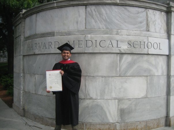 Photograph of a man in his 20s wearing a black graduation cap and gown plus a red hood, he is holding a diploma and standing in front of a marble wall that says "Harvard Medical School". 