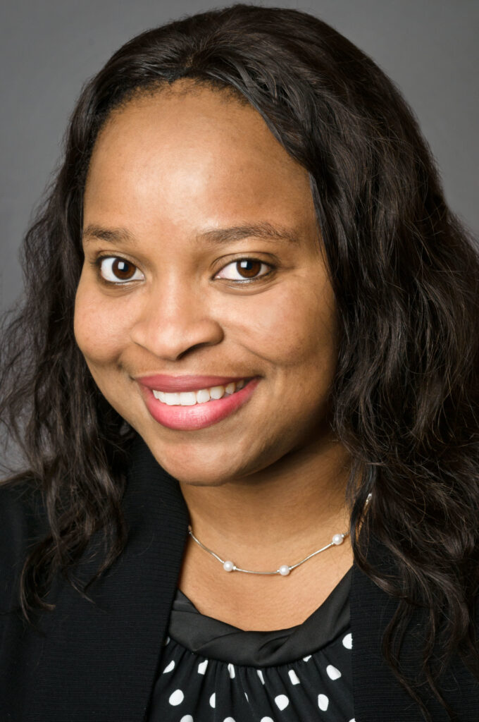 Headshot of a woman in her 20s who has heritage from Nigeria, with medium skin tone and shoulder length black wavy hair. She is wearing a black jacket, black with white polka dot blouse and silver and pearl necklace. She is smiling at the camera. 