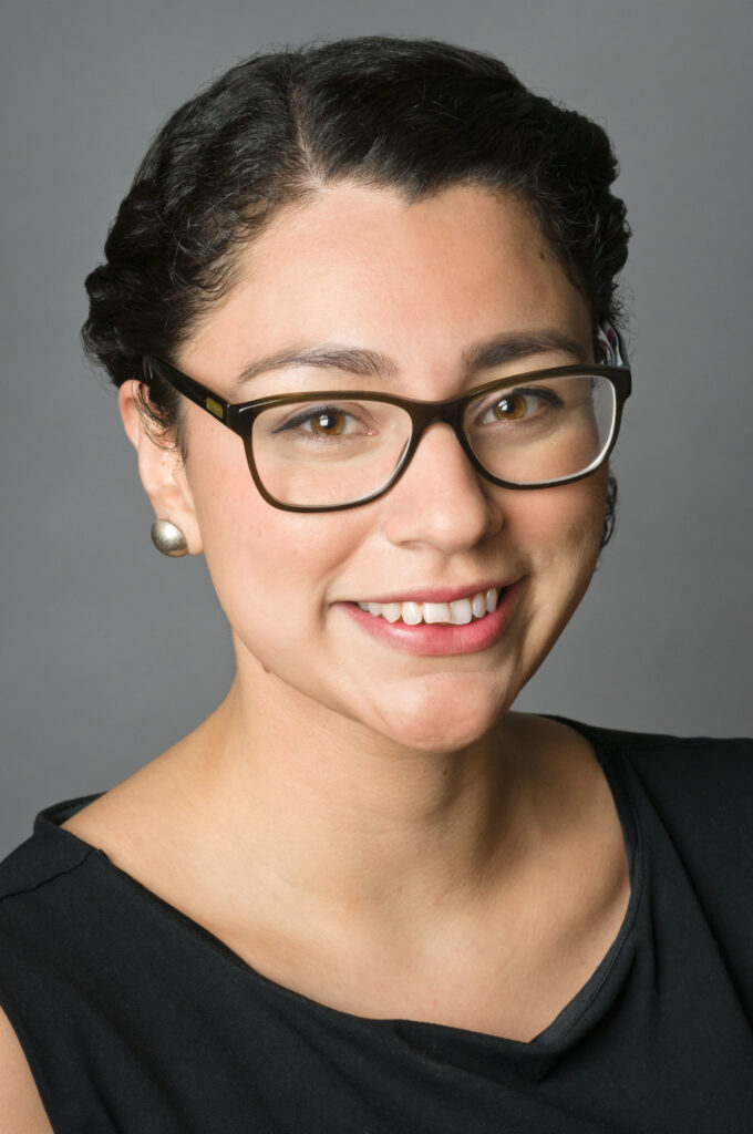 Headshot of a woman in her 20s who has heritage from Honduras and Mexico with light skin tone and black hair, side parted and twisted like a crown and pulled back. She is wearing a black blouse, brass stud earrings and black rimmed glasses. She is smiling at the camera.