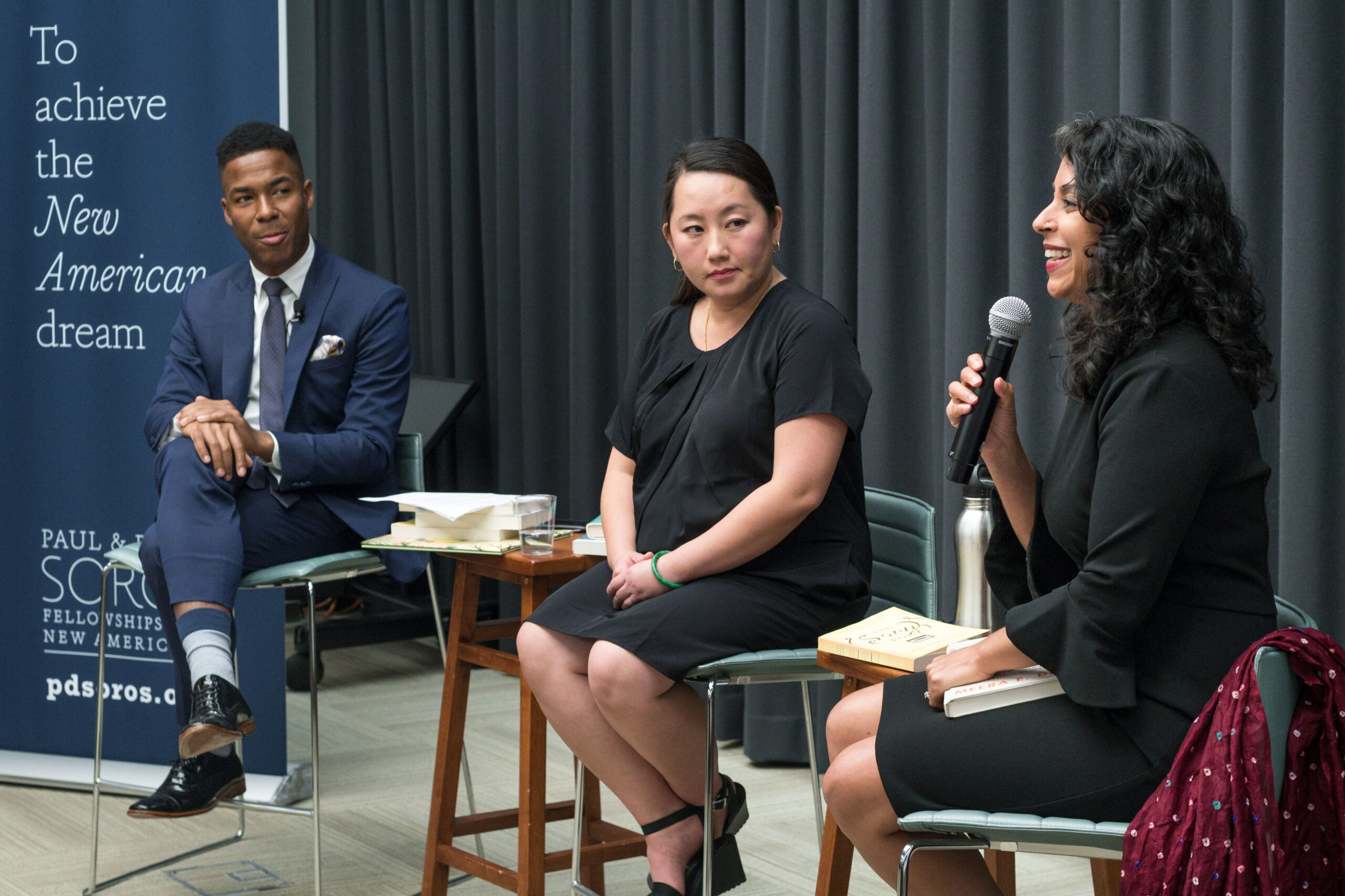 Photograph of three people sitting on stools with a black curtain to the right, they are all looking off camera to the left. The woman on the right has a microphone in hand.