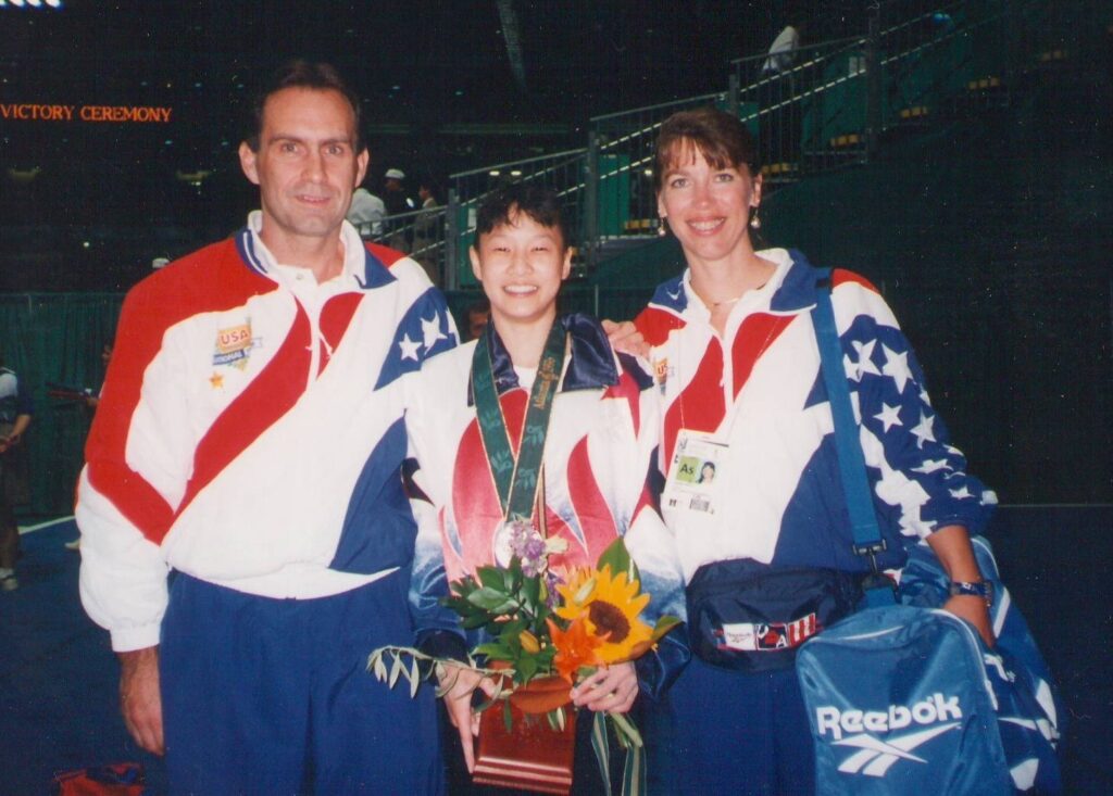 Old photograph of three people wearing matching athletic suits. The young woman in the middle has heritage from Hong Kong and China with light skin tone and black hair pulled back, with bangs. She is wearing a gold medal and holding a bouquet of flowers. 