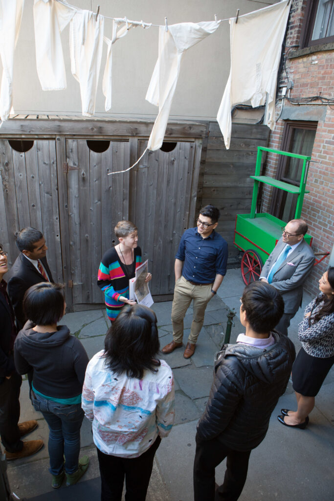 Photograph of a group of people standing in a circle at the Tenement Museum listening to one person talk and point at a piece of paper.