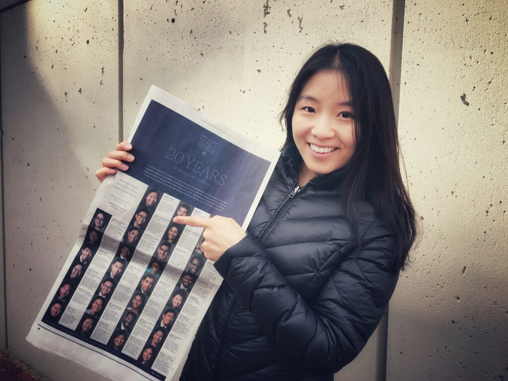 Photograph of a woman in her 20s who has heritage from China with light skin and long straight black hair. She is wearing a black jacket and holding a New York Times.