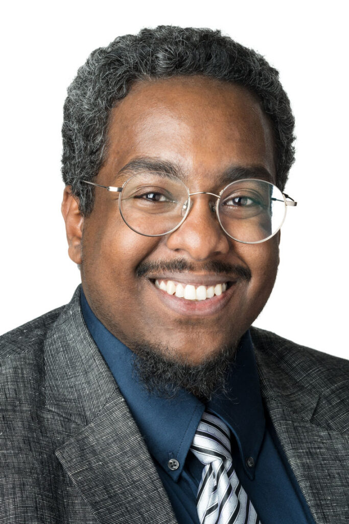 Headshot of a man in his 20s who has heritage from Sudan with medium-dark skin tone, salt and pepper short hair, black mustache and goatee; he is wearing a grey suit, teal button up and white and black tie, he has large round silver thin framed glasses; he is smiling at the camera