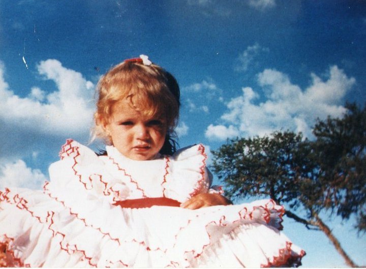 Photograph of a young girl wearing a very frilly white dress with red edging. She is being held up so only the sky and trees can be seen behind her.