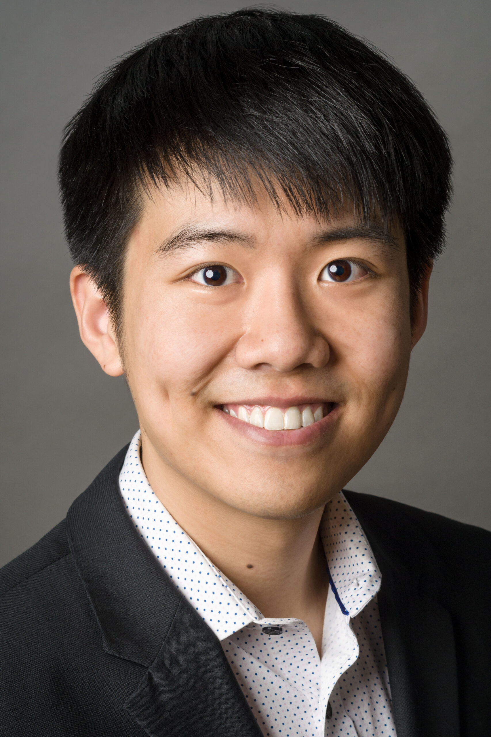 Headshot of a man in his 20s who has heritage from China with light skin tone and black hair. He is wearing a black blazer and a white button up with blue dots. He is smiling at the camera.