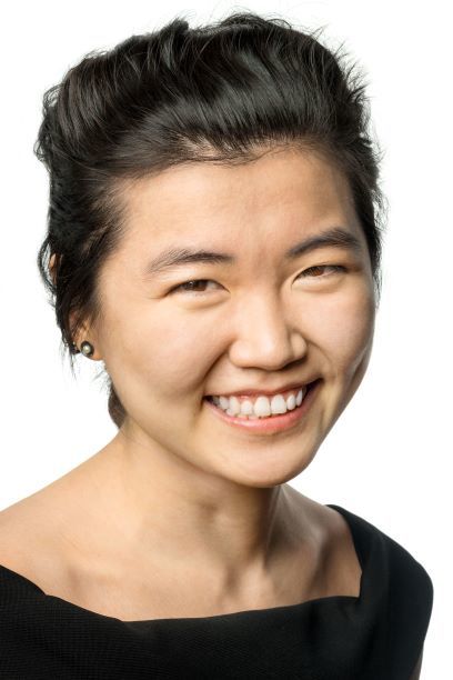 Headshot of a woman in her 20s who has heritage from Jeju Island, South Korea, with light skin tone and black hair that is loosely pulled back. She is wearing a black boat neck top and pearl stud earrings. She is smiling at the camera.