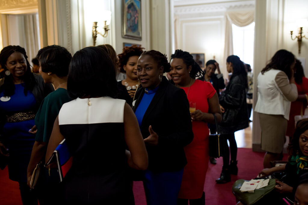 Photograph of a party, many women stand in circles talking. They are all dressed up. Multiple rooms can be seen, they all have very high ceilings, big windows, long curtains and artwork hung on the walls.