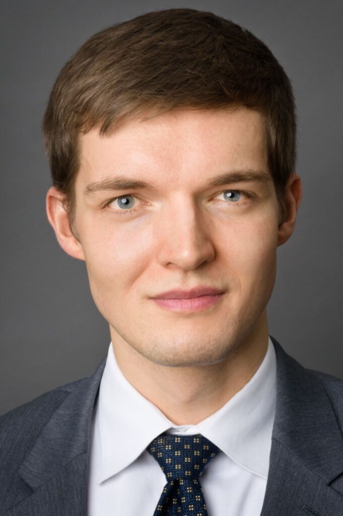 Headshot of a man in his 20s who has heritage from Ukraine with light skin tone and light brown hair. He is wearing a grey suit, white button up shirt and navy tie. He is looking at the camera.