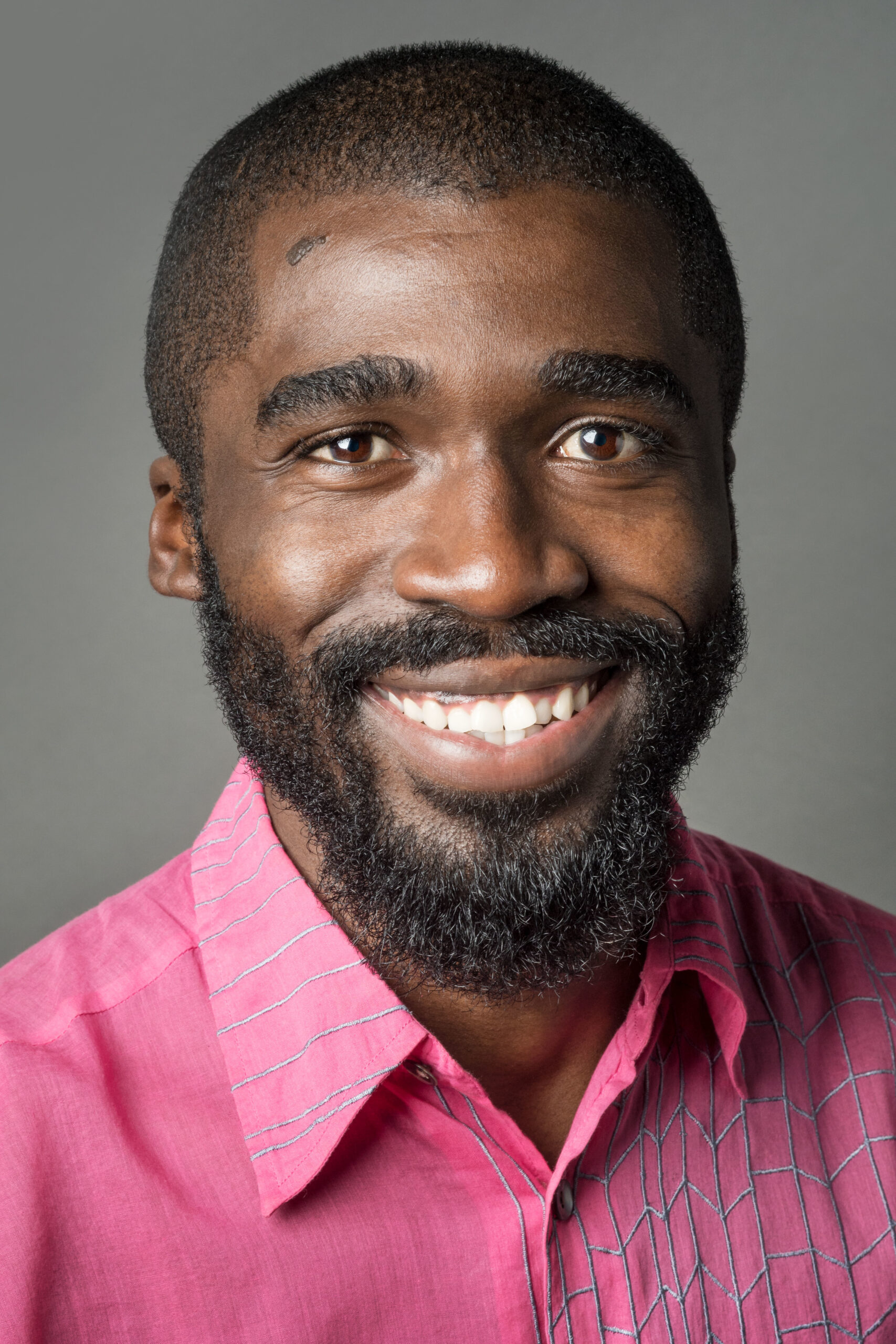 Headshot of a man in his 20s who has heritage from Guyana with dark skin tone, buzzed black hair and a beard. He is wearing a bright pink button up shirt with grey embroidered accents on the collar and left side. He is smiling at the camera.