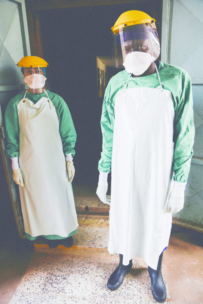 Photograph of two people standing at a doorway in full PPE (coverall, apron, rubber boots, gloves, N95 masks, face shields and hair coverings. 