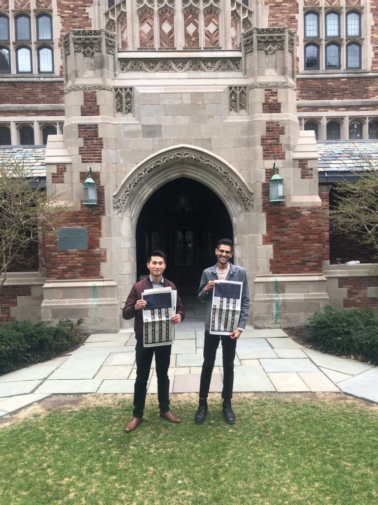 Photograph of two men in their 20s standing in front of an intricately decorated doorway to a stone and brick building. They are both holding a New York Times.