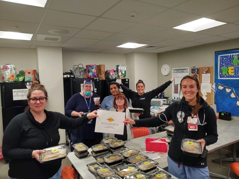 Photograph of six people standing around a table that has to go containers of food. They are holding a piece of paper and looking at the camera.