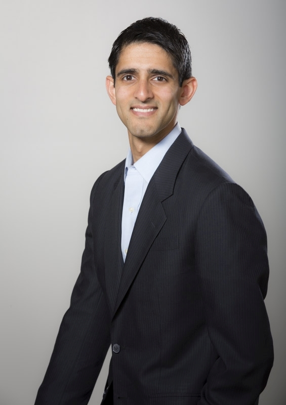 Headshot of a man in his 30s who has heritage from India with light skin tone and short black hair. He is wearing a black pinstriped suit, light blue button up shirt. 