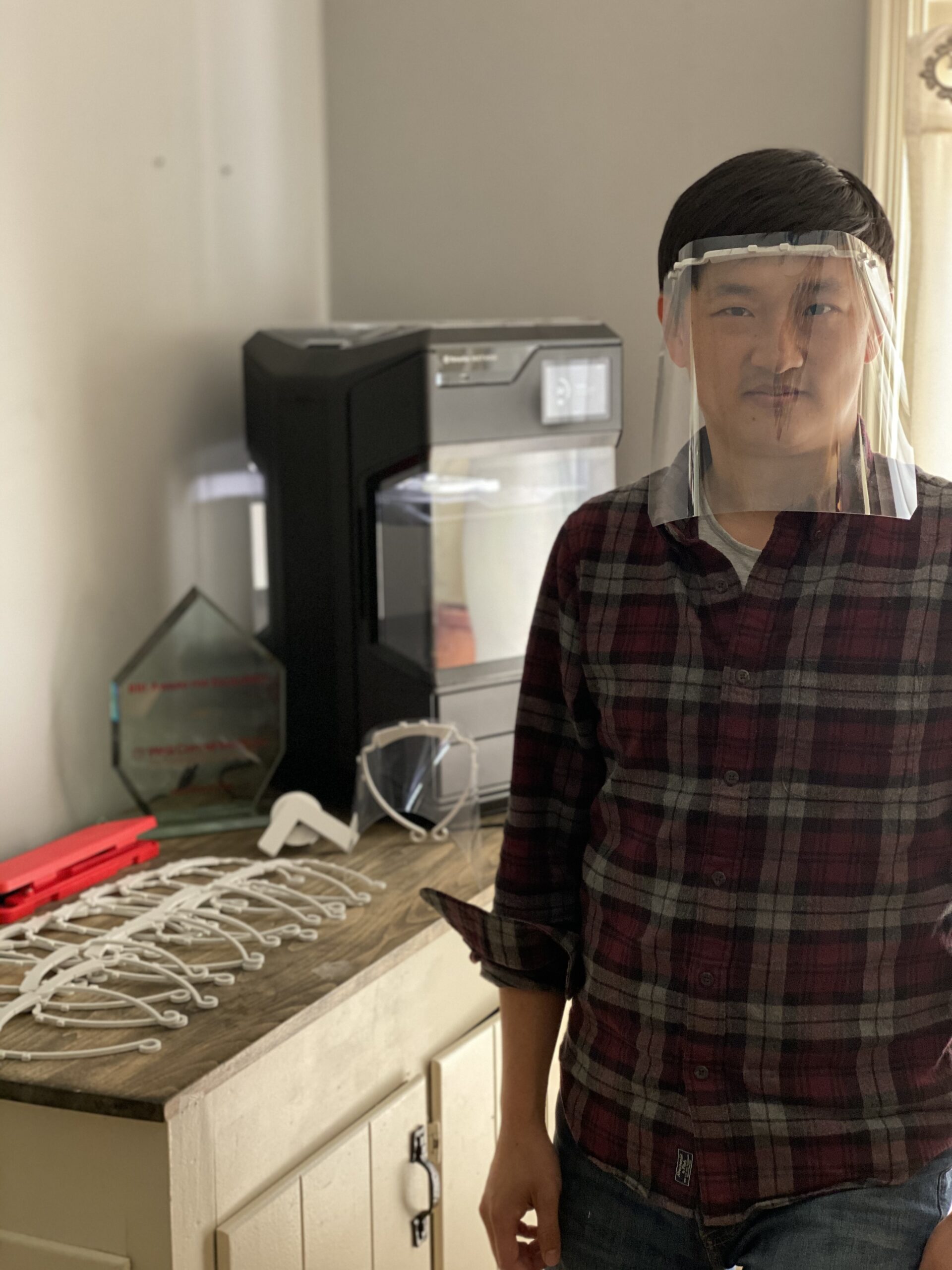 Photograph of a man in his 20s with heritage from China, he has light skin tone and black hair. He is wearing a clear plastic face shield, red flannel shirt and jeans. He is standing next to a table that has the hard wear to make face shields.