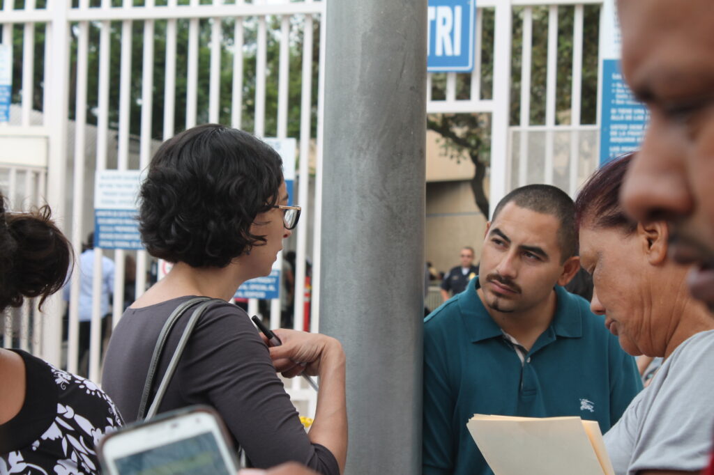 Photograph of two people talking. The woman on the left who has heritage from Iran has light skin tone and short black curly hair. She is wearing a grey dress and gold and black glasses. She is talking to a man wearing a teal polo. 