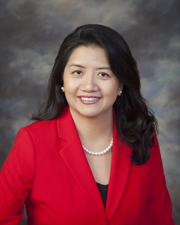 Headshot of a woman in her 40s who has heritage from Viet Nam with light skin tone and long black curled hair. She is wearing a red blazer, black blouse, pearl necklace and pearl stud earrings. She is smiling at the camera. 