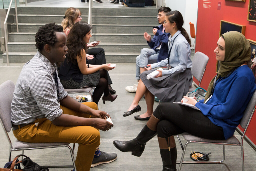 Photograph of six people sitting in chairs, facing one another making three groups of two. They appear to be in discussion. They are sitting in a room with a red wall and gallery style photos on the wall. 