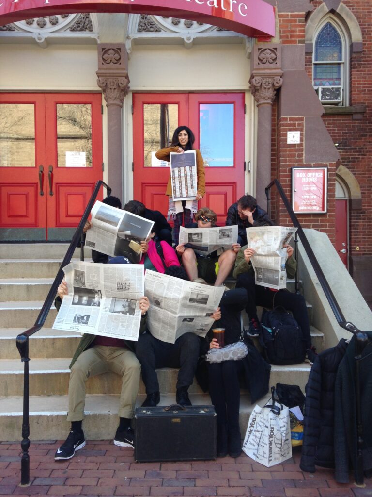 Photograph of a woman in her 20s with light skin tone and long wavy black hair, stands at the top of a staircase holding the New York Times. In front of her the steps are filled with people sitting with open New York Times pages. 