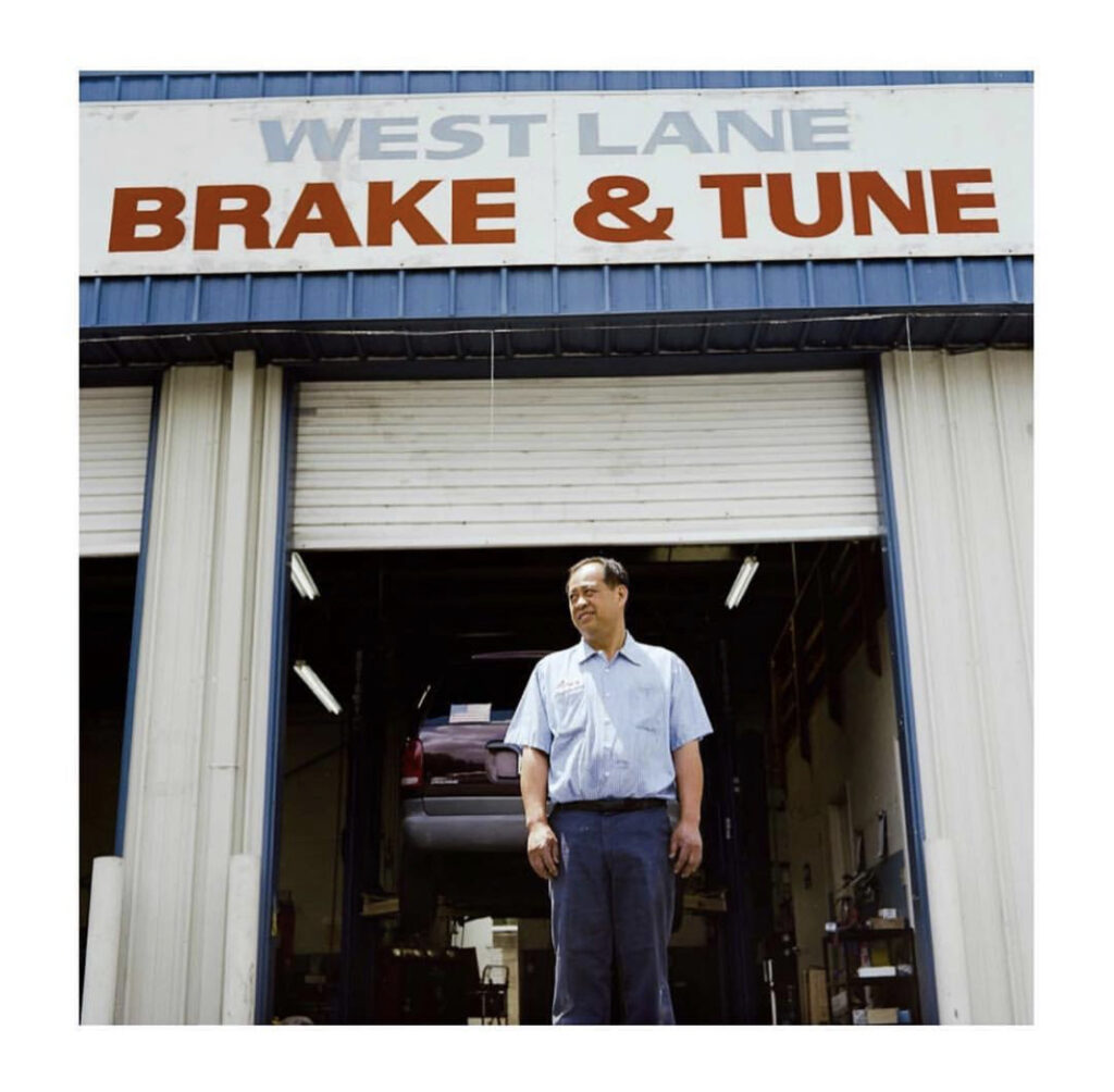 Old photograph of a man in his 40s wearing navy blue pants and a light blue short sleeve button up. He is standing in front of a garage door, above the door a sign that says "West Lake Brake & Tune". 