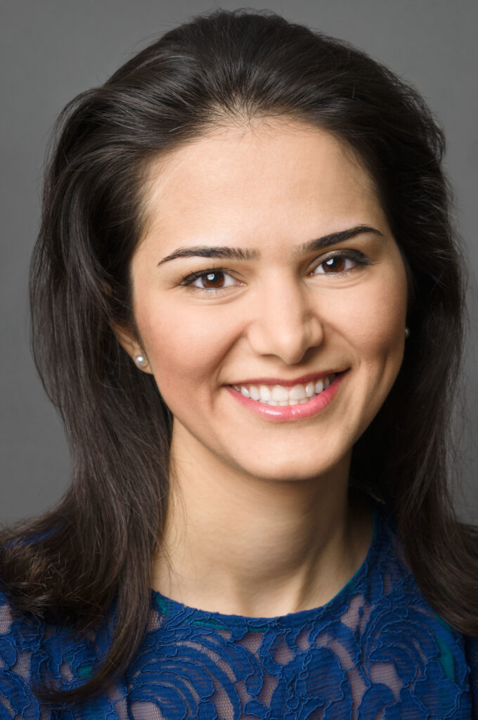 Headshot of a woman in her 20s who has heritage from Iran with light skin tone and combed back long brown hair. She is wearing a blue lace blouse and pearl stud earrings. She is smiling at the camera.