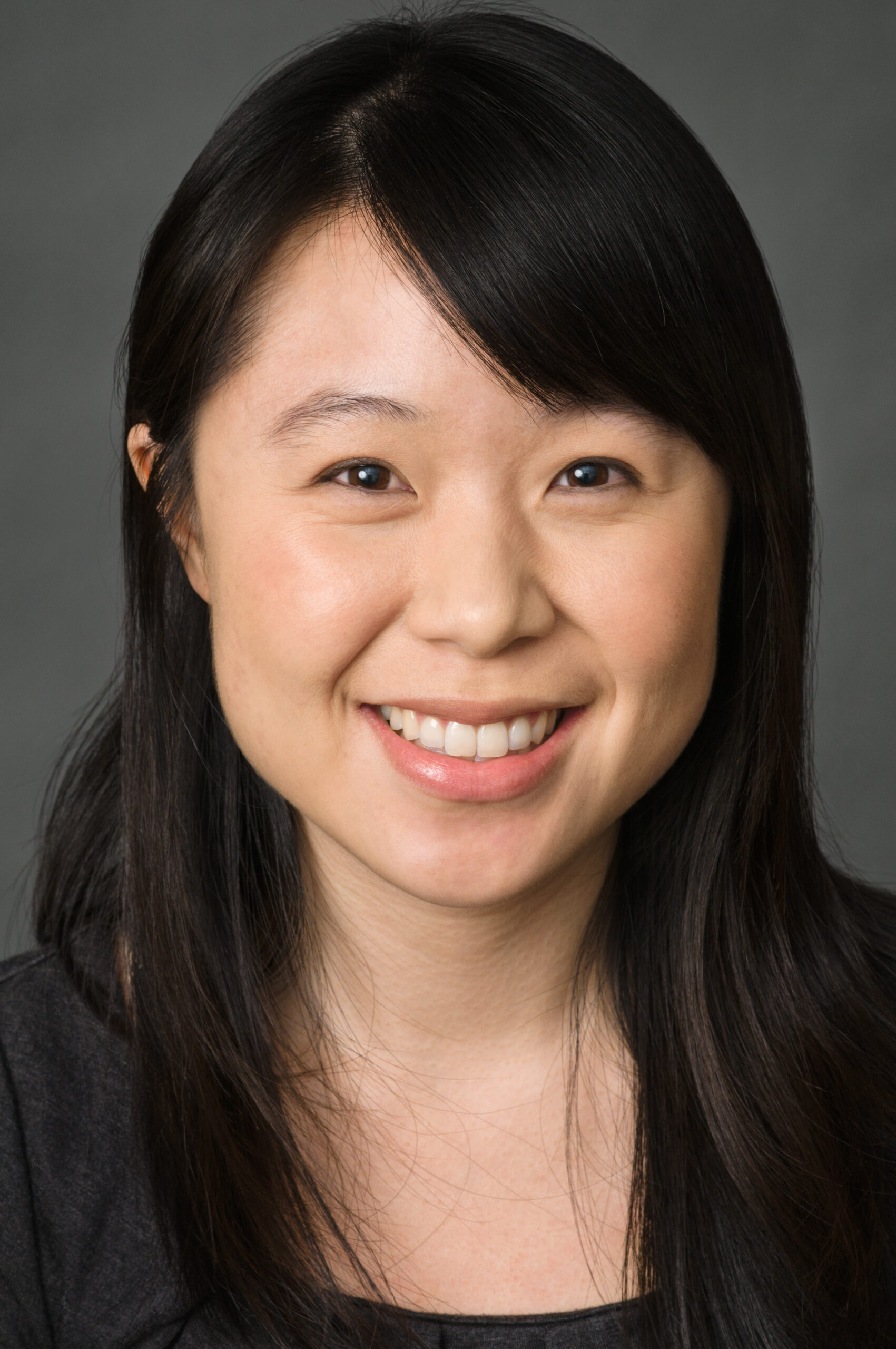 Headshot of a woman in her 20s who has heritage from China with light skin tone and long black straight hair with side bangs, across her left forehead. She is wearing a black top and smiling at the camera. 