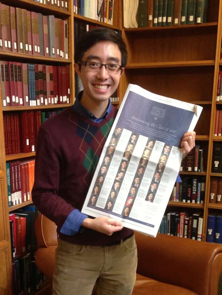 Photograph of a man in his 20s with light skin tone and dark brown hair. He is wearing an argyle maroon, black and green sweater, with a blue button up underneath and holding up a New York Times. He is standing in a corner of bookshelves, that have many books on them. 