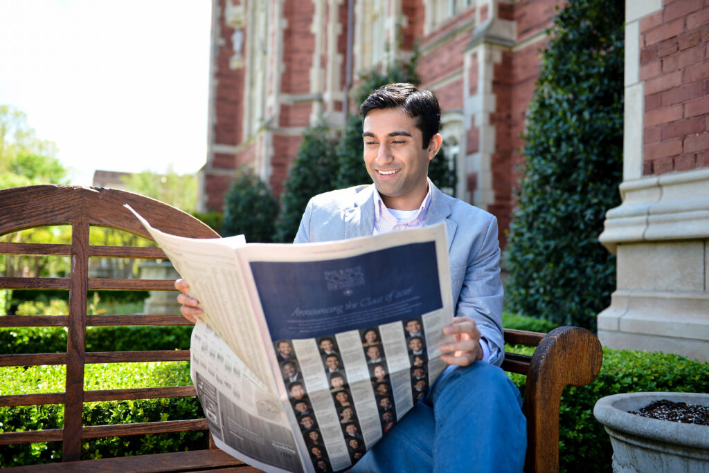 Photograph of a man in his 20s who has heritage from United Kingdom with light-medium skin tone and black hair, he is wearing a grey suit, sitting on a bench appearing as if he is reading the New York Times, a red brick building can be seen behind him. 