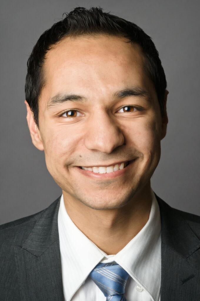 Headshot of a man in his 20s with heritage from United Arab Emirates, he has medium-light skin tone and short black hair. He is wearing a dark grey suit, white button up shirt and blue tie. He is smiling at the camera.