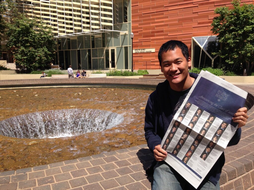 Photograph of a man in his 20s with medium skin tone and short black hair. He is wearing a long sleeve shirt, jeans and holding a New York Times. 