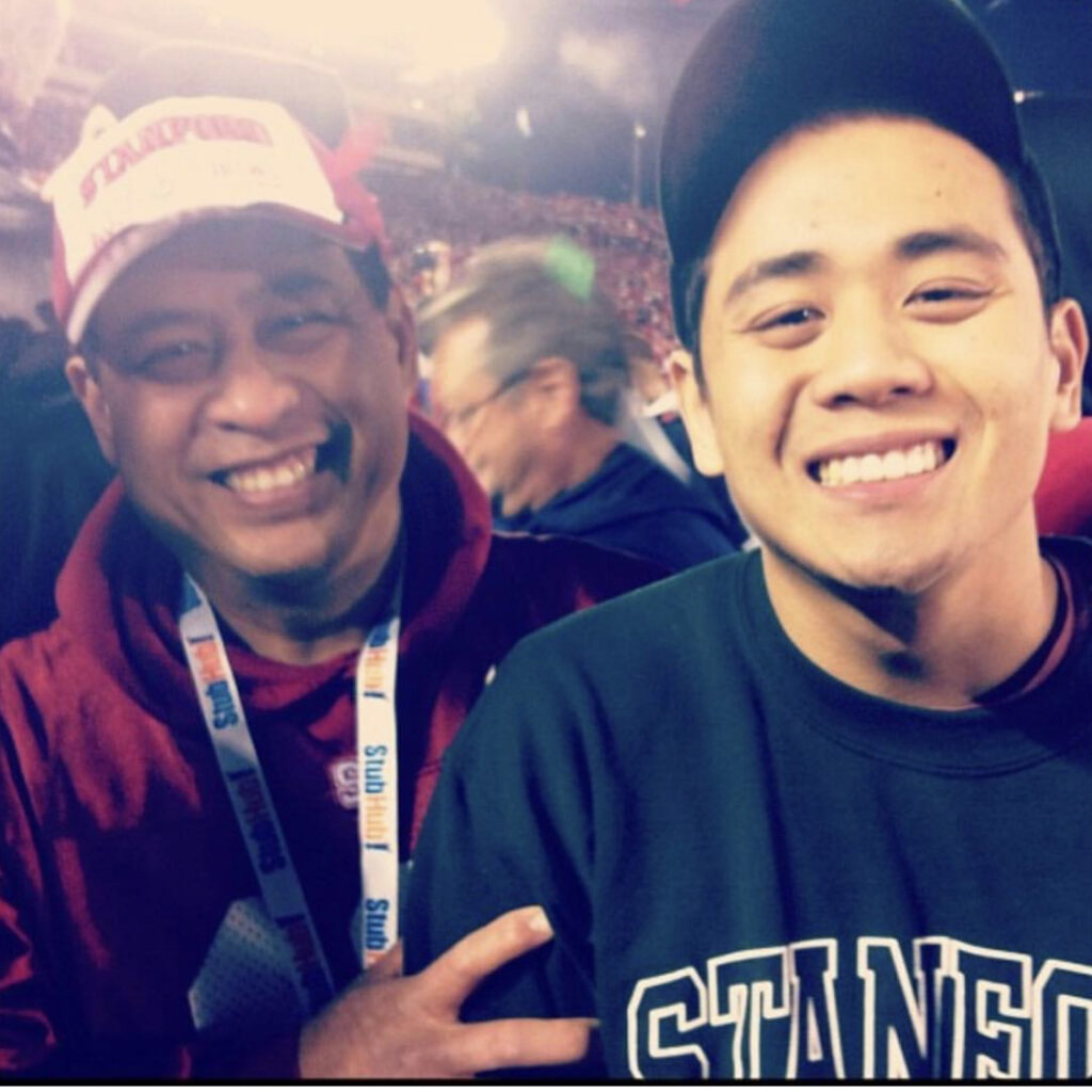 Photograph of two men, one in his 40sish and one an older teenager. A large crowd can be seen behind them. They are both smiling big at the camera. 