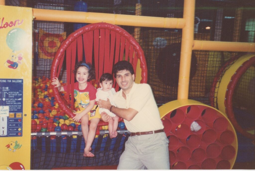 Old photograph of a dad and two kids. The dad has light skin tone and black hair, he is wearing a white polo and grey slacks. He is standing with the kids at the entrance to a ball pit.