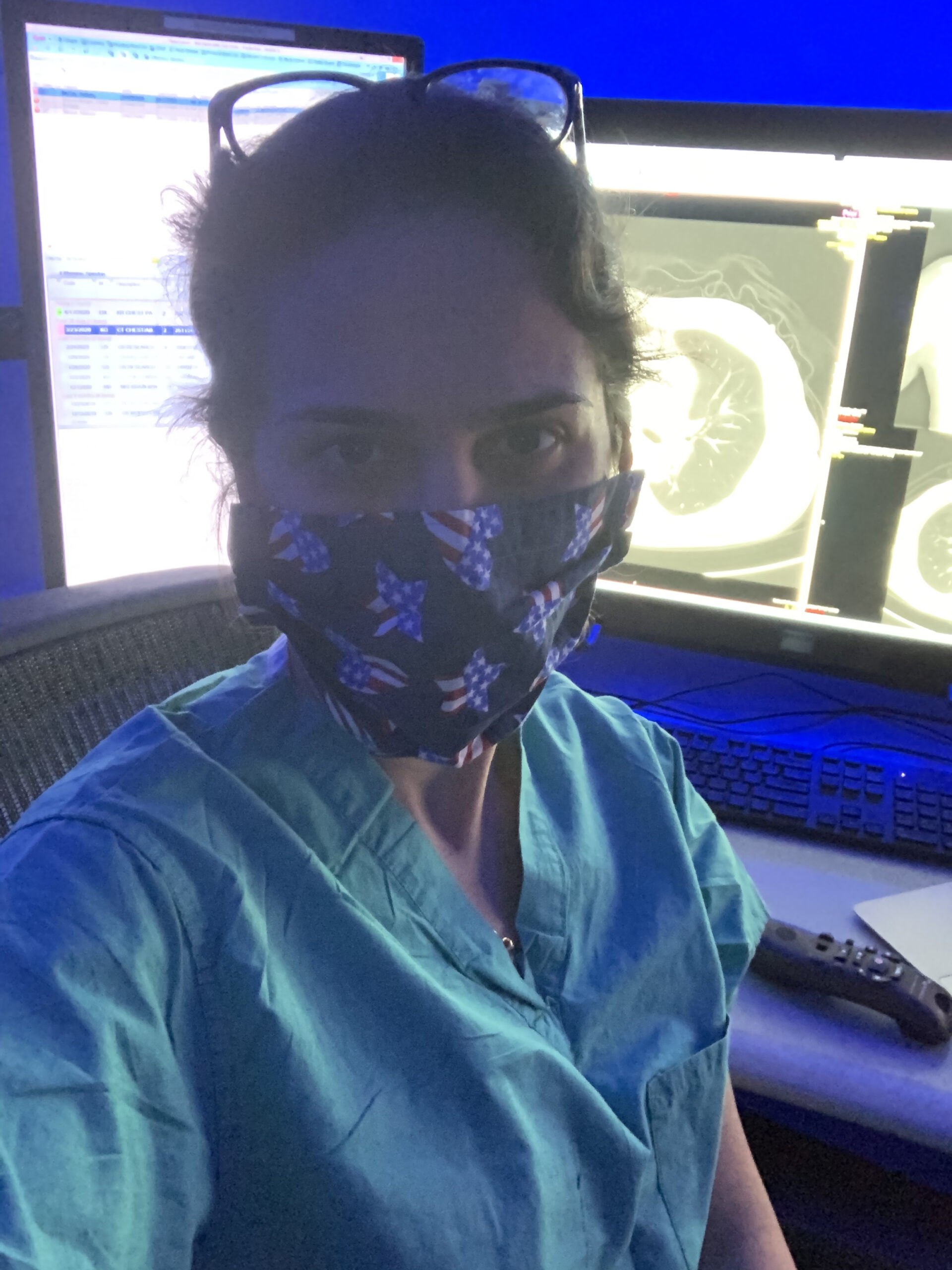 Selfie of a woman in her 30s who has heritage from Iran with light skin tone and brown hair pulled back. She is wearing blue scrubs and an American flag star mask with glasses on her head. She is sitting in front of medical scans on screens.