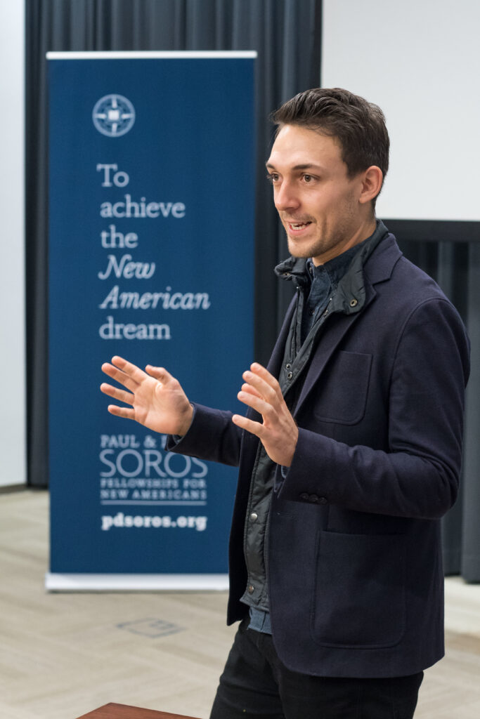 Photograph of a man speaking, his arms up in gesture, a screen can be seen behind him. A Paul & Daisy Soros Fellowships banner is behind him. 