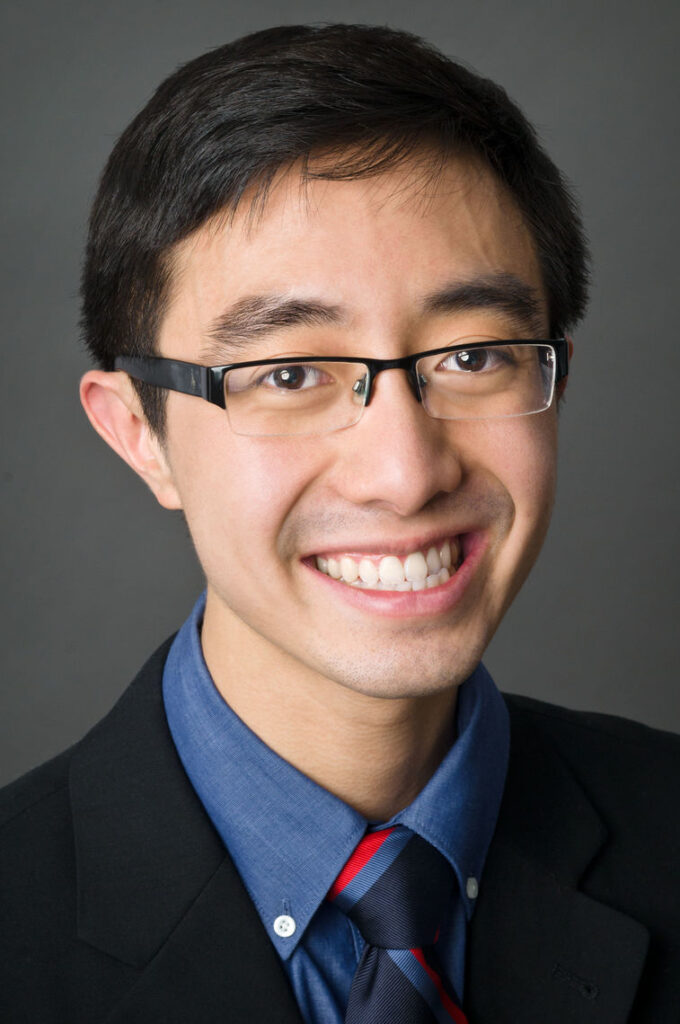 Headshot of a man in his 20s with heritage from Vietnam with light skin tone and black short hair; he is wearing a black blazer and blue button up shirt, he wears half-rimmed thin rectangle glasses and smiles at the camera