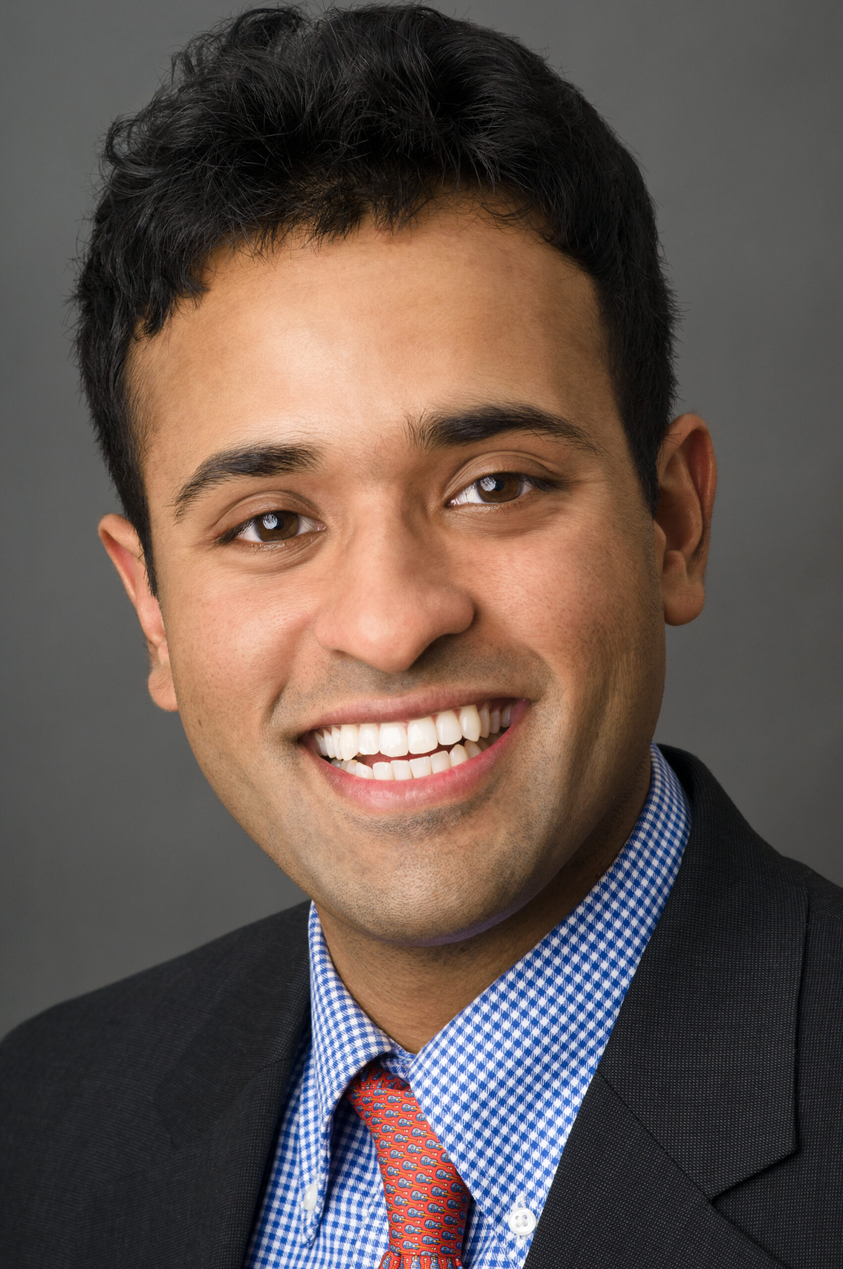 Headshot of a man in his 20s who has heritage from India with medium skin tone and crew cut black hair. He is wearing a black suit, blue and white gingham button up and red tie. He is smiling at the camera.