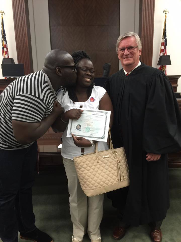 Photograph of three people. The woman in the middle has dark skin tone and black long braided hair, she is holding a certificate. The man to her right also has dark skin tone and is bald, he is leaning over to give her a kiss on the cheek. On the right side of the photo a man in his 60s with light skin tone and grey hair wearing judge's robes stands smiling at the camera. 