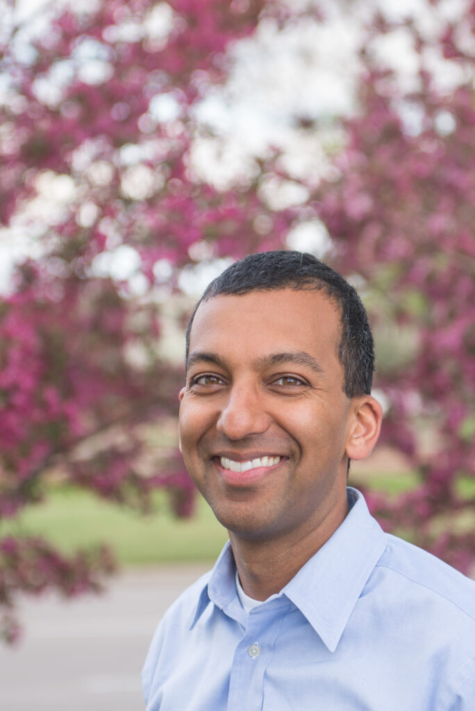 Headshot of a man in his 40s who has heritage from India with medium skin tone and buzzed black hair. He is wearing a blue button up shirt and smiling at the camera. Pink blossom trees can be seen behind him. 