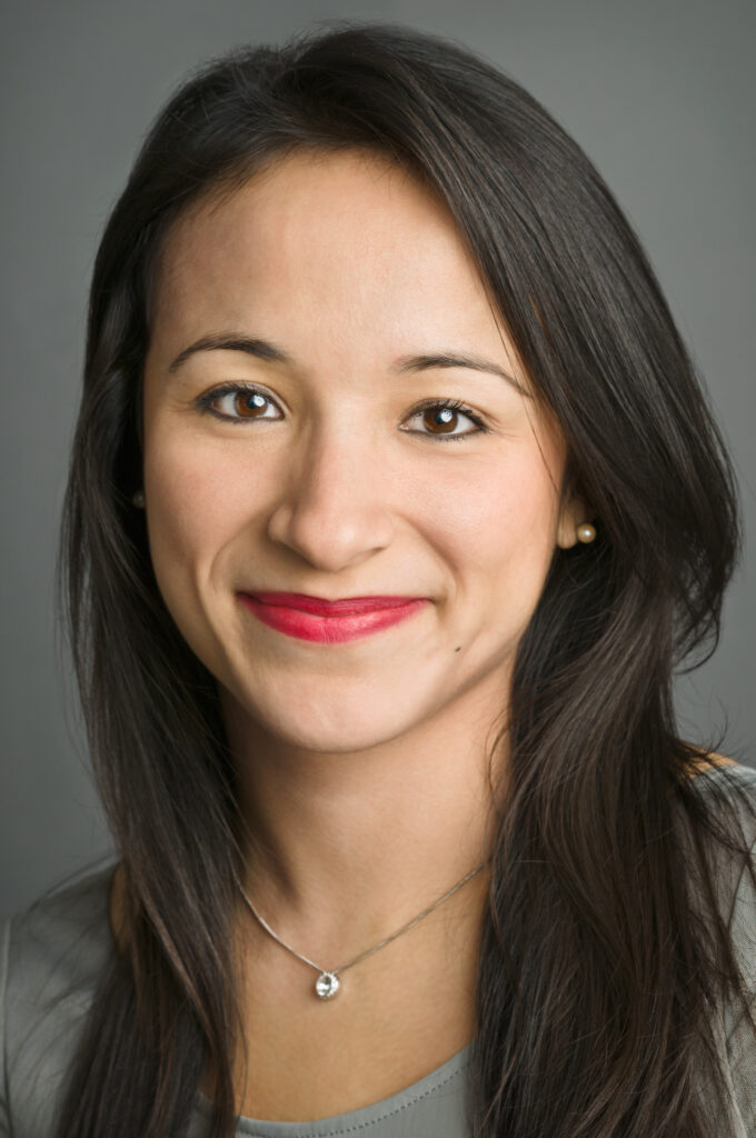 Headshot of a woman in her 20s who has heritage from Ecuador and Poland, with light skin tone and long straight black hair. She is wearing a grey top, silver necklace and pearl stud earrings. She is smiling at the camera. 