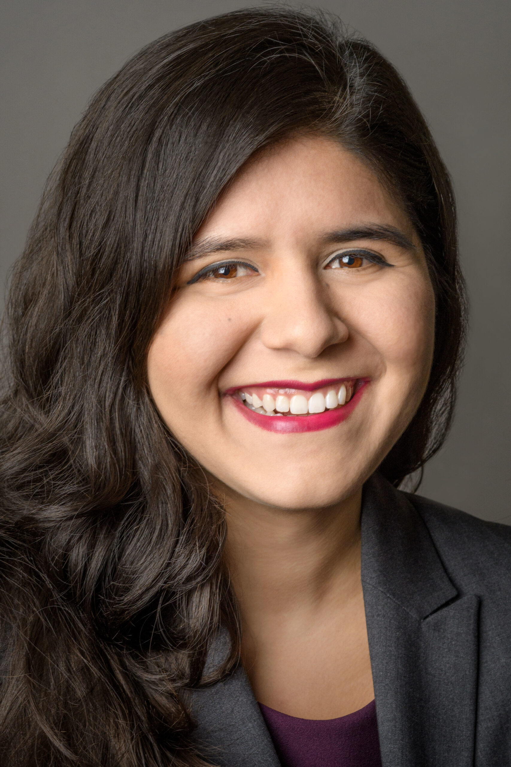 Photograph of a woman in her 20s who has heritage from Mexico with light-medium skin tone and long dark brown curled hair. She is wearing a dark brown blazer and purple blouse, she is looking at the camera and smiling. 