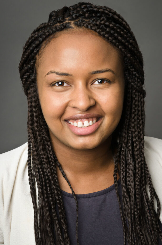 Headshot of a woman in her 20s who has heritage from Somalia with medium skin tone and long black braided hair. She is wearing a white jacket, and navy blouse. She is smiling at the camera.