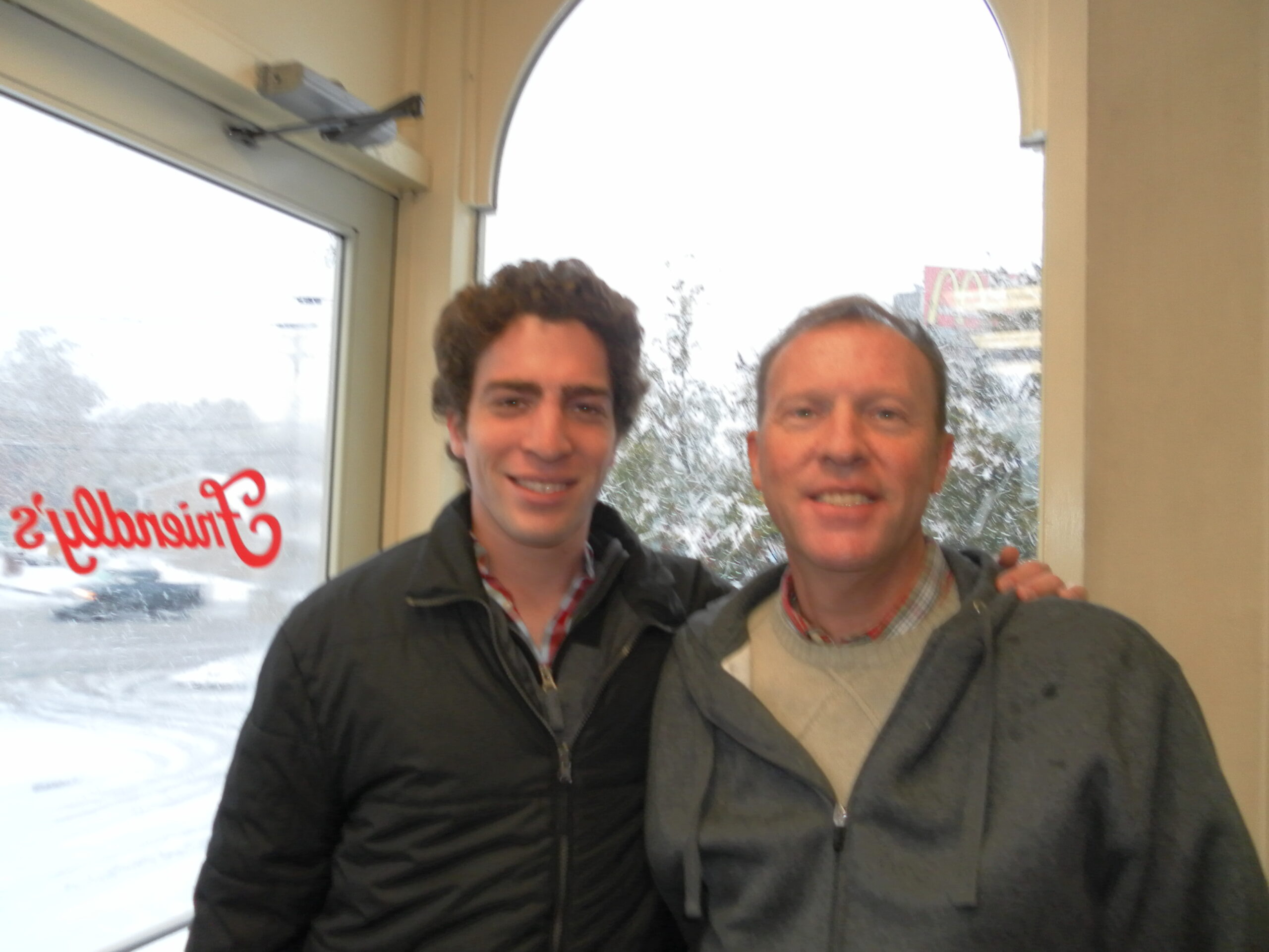 Photograph of a teenager who has heritage from Brazil with light skin tone and auburn curly short hair. He is wearing a winter coat and has his arm around the man in the photo, who is in his 50s with light skin tone and short hair. 