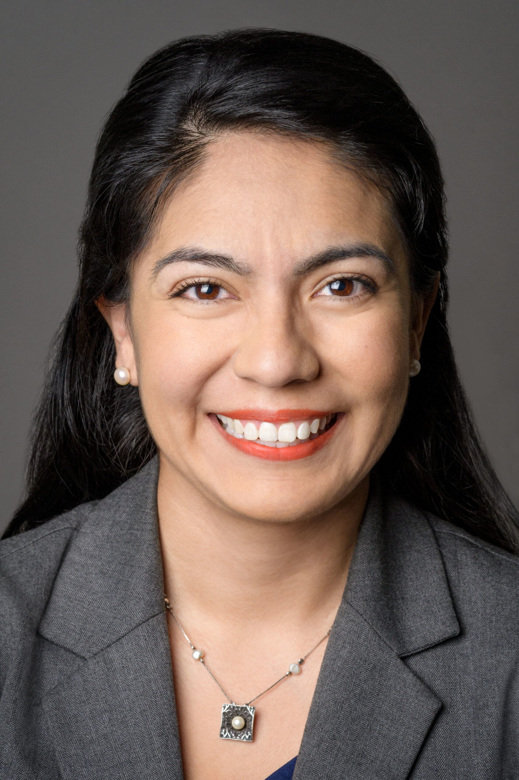 Headshot of a woman in her 20s who has heritage from Mexico with light skin tone and long black straight hair, half pulled back. She is wearing a grey blazer, a silver and pearl necklace and pearl stud earrings. She is smiling at the camera.