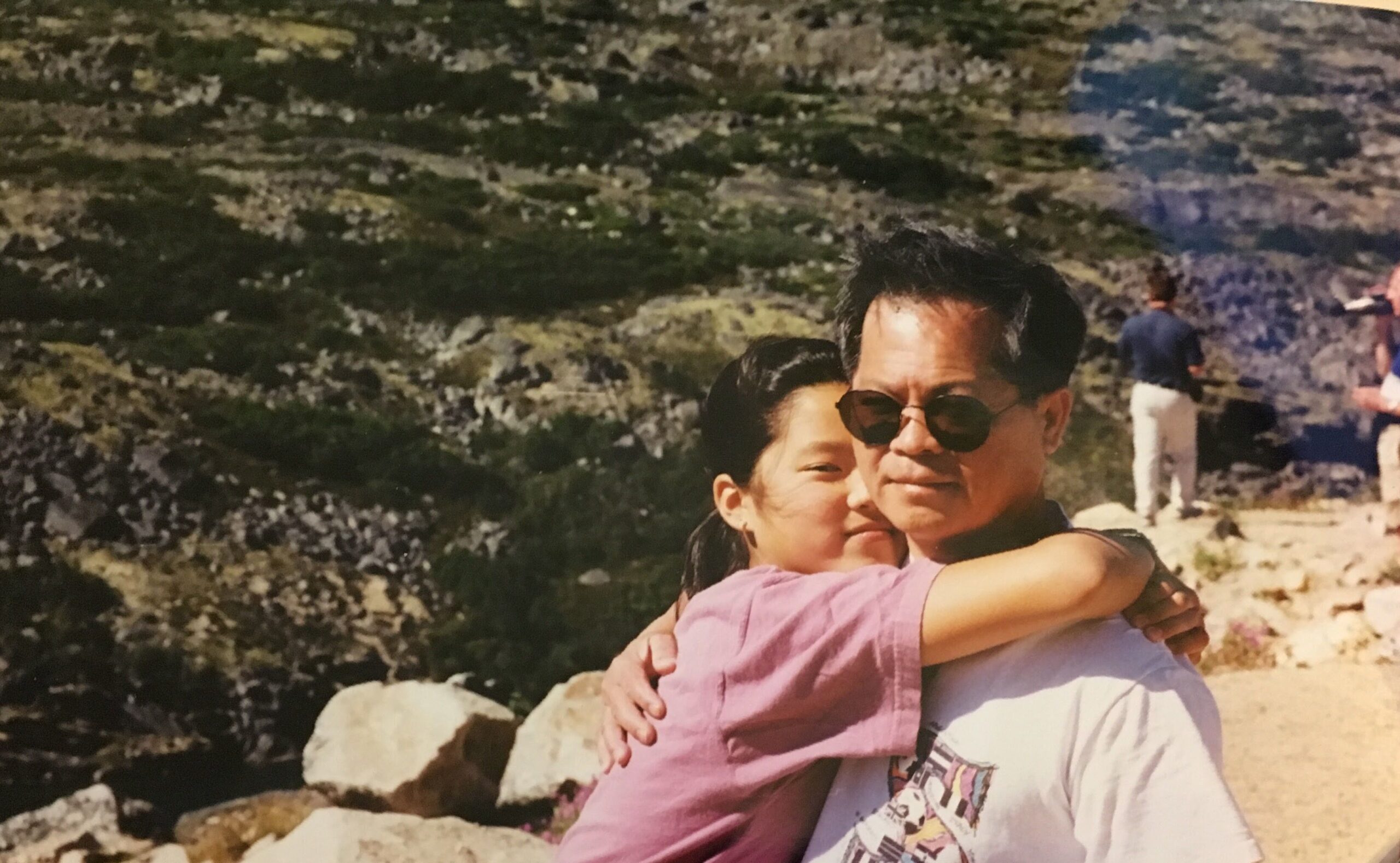 Old photograph of a woman in her 20s who has heritage from China and Taiwan with light skin tone and black hair, hugs a man around the neck. They are both looking at the camera, he has on round sunglasses. Green grass and rocky landscape can be seen behind them. 