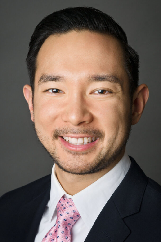 Headshot of a man in his 20s who has heritage from Viet Nam and Japan, with light skin tone and combed back black hair. He is wearing a black suit, white button up shirt and pink tie. He is smiling at the camera.