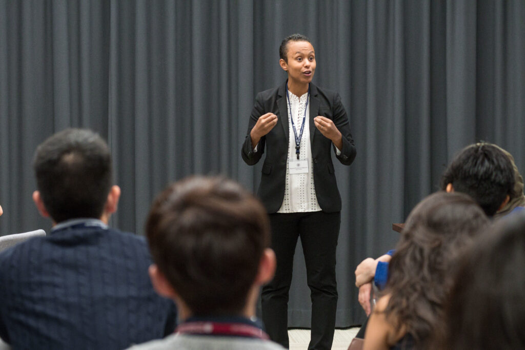 A woman in her 30s with medium skin tone and tightly pulled back black hair, wearing a black blazer, white blouse and black pants. She is standing in front of a grey curtain and speaking, gesturing with her hands. 