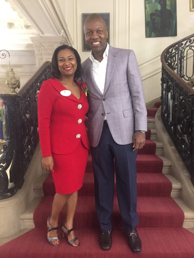 Photograph of a woman in her 40s who has heritage from Haiti with dark skin tone and chin length black hair. She is wearing a red skirt suit with a nametag on her right lapel and a rose on her right lapel and strappy heels. Next to her a man with dark skin tone wearing a grey jacket, white button up shirt and navy pants. They are standing in front of a staircase. 